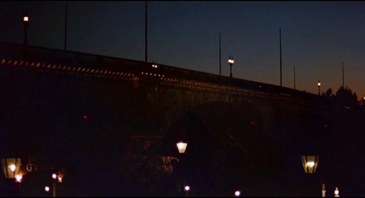 Olivia (1983) movie still: establishing — A stone arch bridge spans across a dark expanse at twilight under a clear sky. A series of evenly spaced…