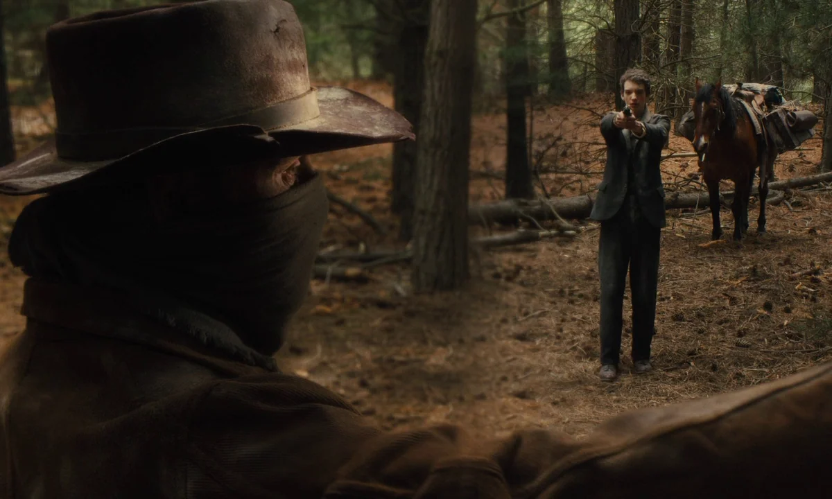 Slow West (2015) movie still: medium — A dense pine forest floor covered in needles and fallen tree trunks. A young man stands in the distance…