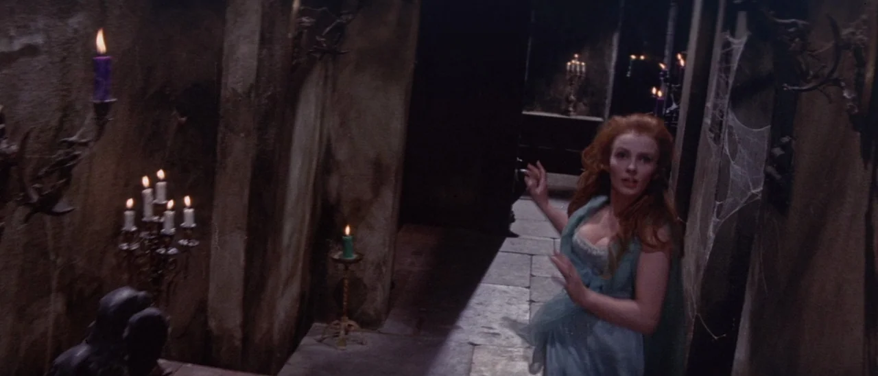 The Tomb of Ligeia (1964) movie still: medium — A dark stone corridor features lit candles on wall sconces and draped spider webs. A young woman…