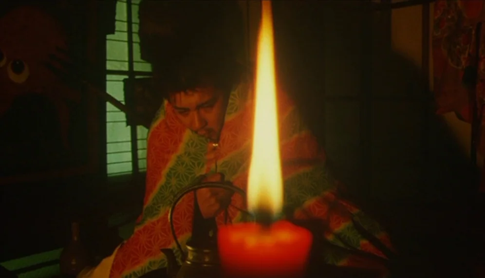Gozu (2003) movie still: medium — A dimly lit traditional Japanese room with a paper sliding door in the background. A man sits at a table draped in…