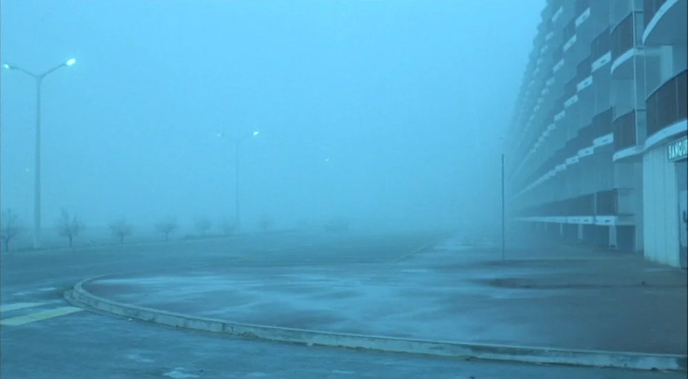 Un Flic (1972) movie still: establishing — A vast, empty parking lot stretches out under heavy, thick blue fog. A modern multi-story apartment block…