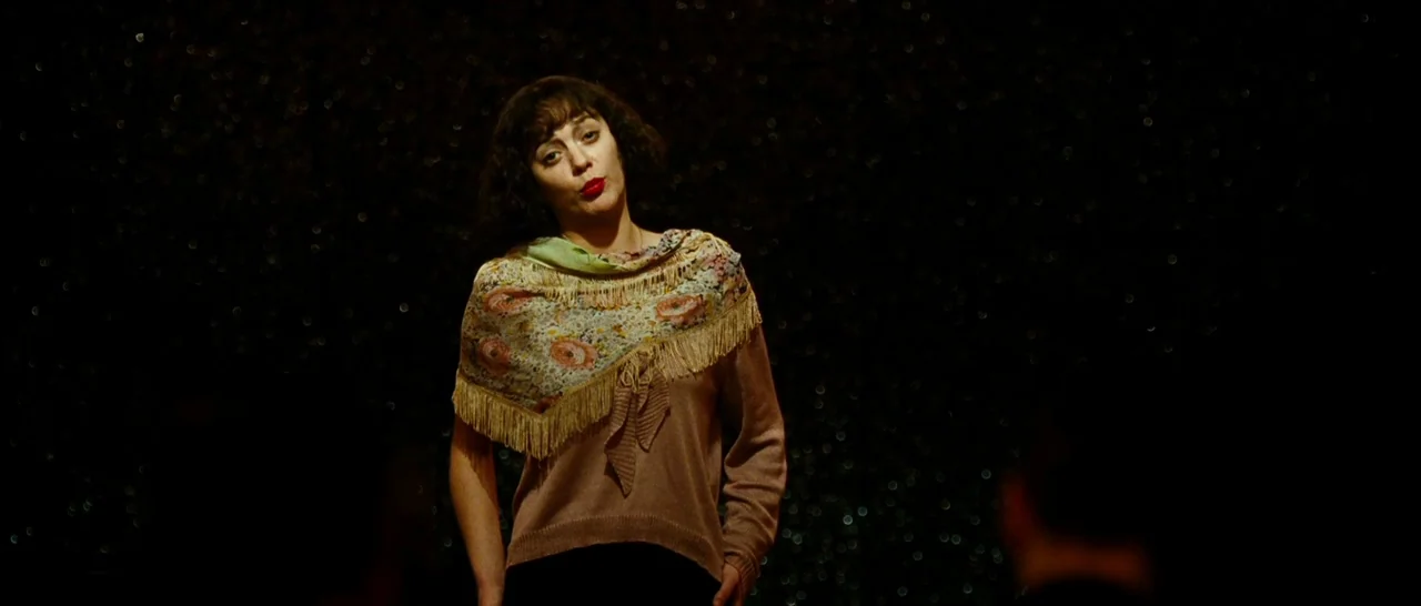 La Vie En Rose (2007) movie still: medium — A dark stage features a performer standing in front of a glittery backdrop. The woman wearing a floral…