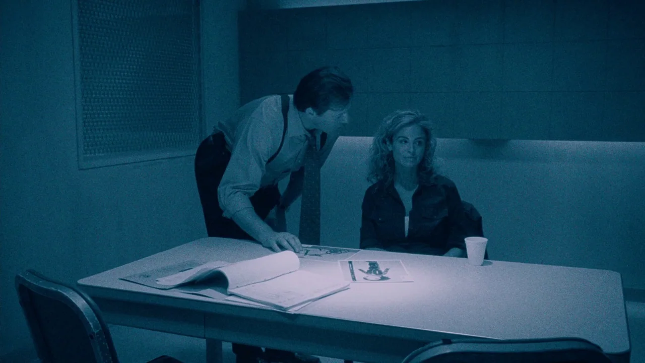 Saw IV movie still: medium — An interrogation room features a metal table, chairs, and plain walls under blue light. A man leaning over the table…