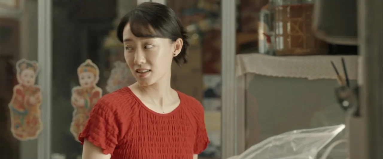 Yolo (2024) movie still: medium — A cluttered shop interior features glass shelving units and paper decorations on walls. A woman in a red shirt…