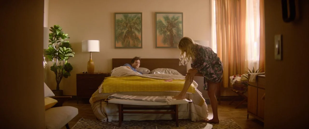 Palm Springs (2020) movie still: wide — A sunlit room features a made bed with a yellow throw blanket and two framed pictures of palm trees. A woman…