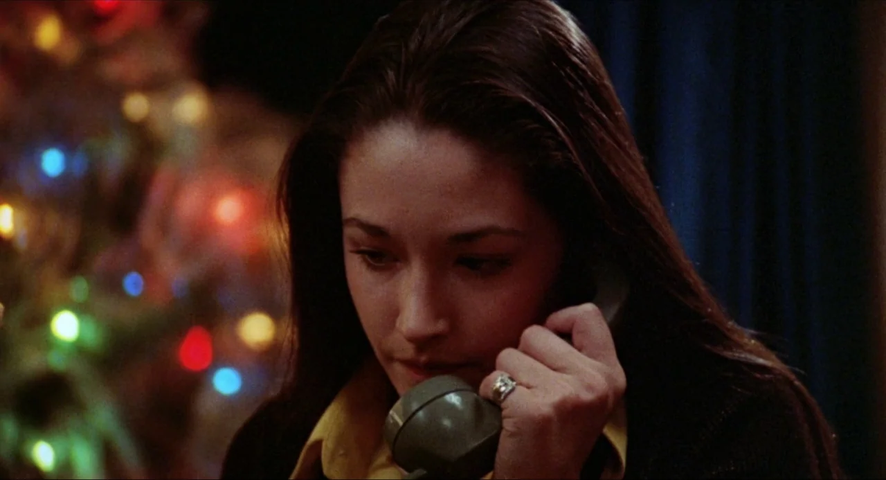 Black Christmas (1974) movie still: closeup — A dark indoor space with a blurred Christmas tree in the background. A young woman holds a rotary…