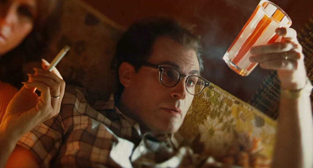 A Serious Man (2009) movie still: closeup — A dimly lit room features patterned furniture and warm, dated interior lighting. A man wearing glasses…