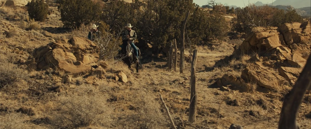 Frontera (2014) movie still: wide — A dry, arid desert landscape features rugged rock formations and sparse scrub brush. Two men in cowboy hats ride…