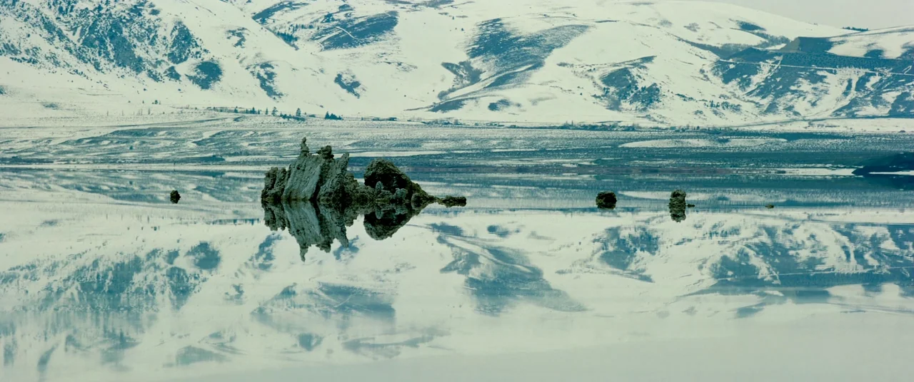 Samsara (2011) movie still: establishing — A vast, calm lake reflects snow-covered mountains and jagged rocky tufa formations. The water surface…