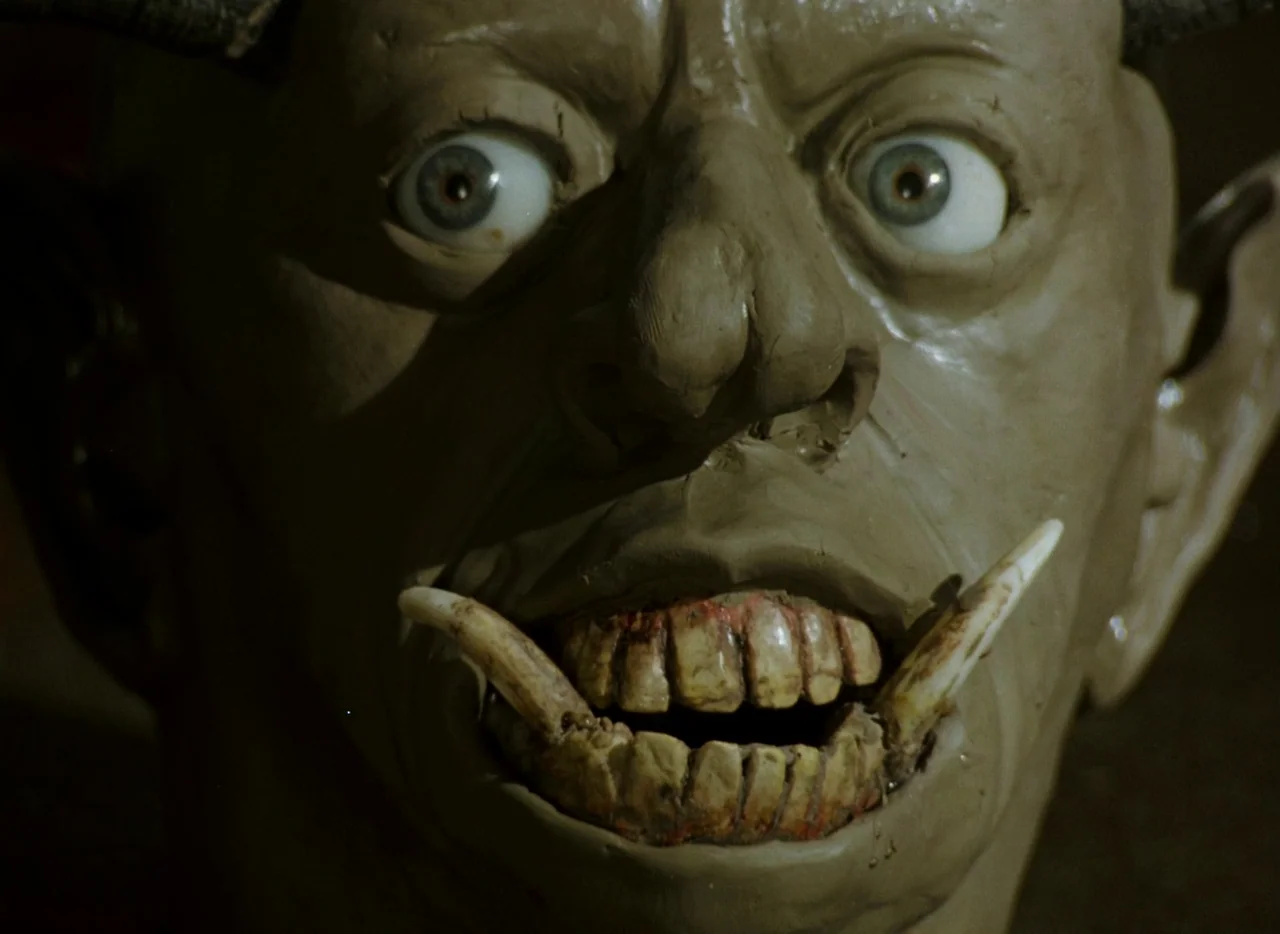 Faust (1994) movie still: closeup — A close-up of a sculpted clay monster face with human eyes and sharp tusks. The creature maintains a fixed…