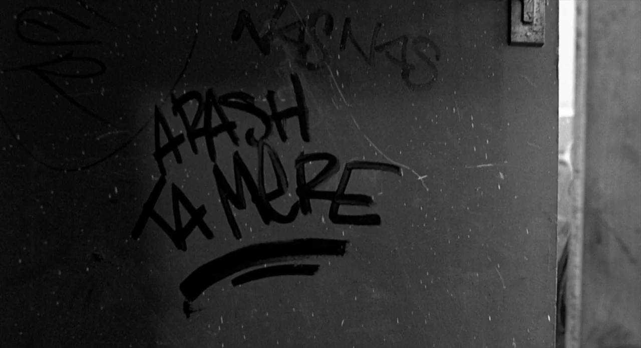 La Haine (1995) movie still: closeup — A flat door surface marked with handwritten black marker graffiti reading Arash and Ta Mere. The door is…