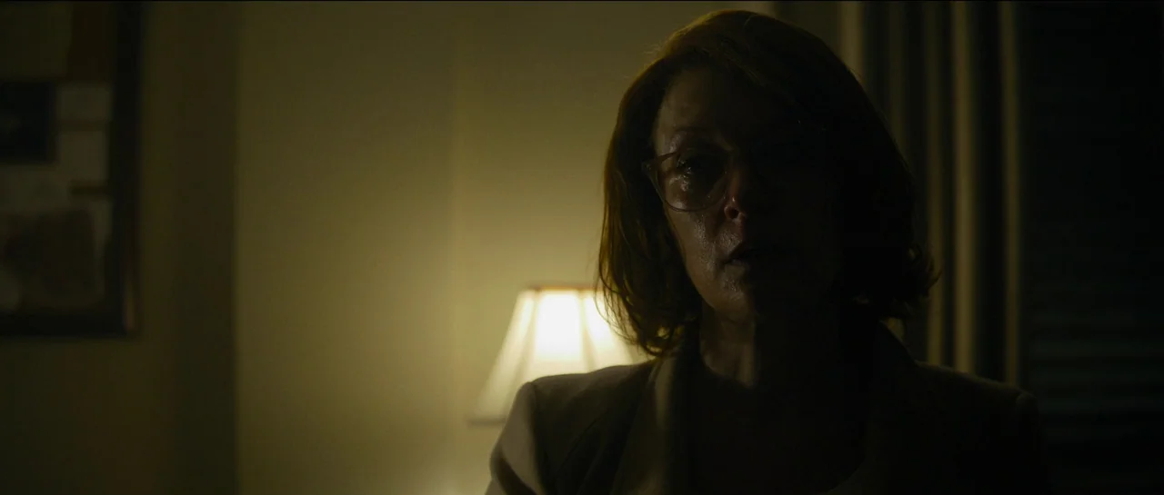 The Killer (2023) movie still: closeup — A dimly lit room interior features a woman in the foreground. She stands near an illuminated lampshade…