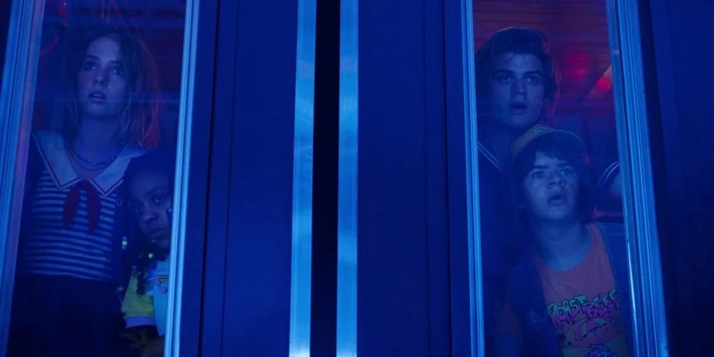 Stranger Things (2019) movie still: medium — Four teenagers stand inside a small, dimly lit structure looking through glass panels. The interior is…