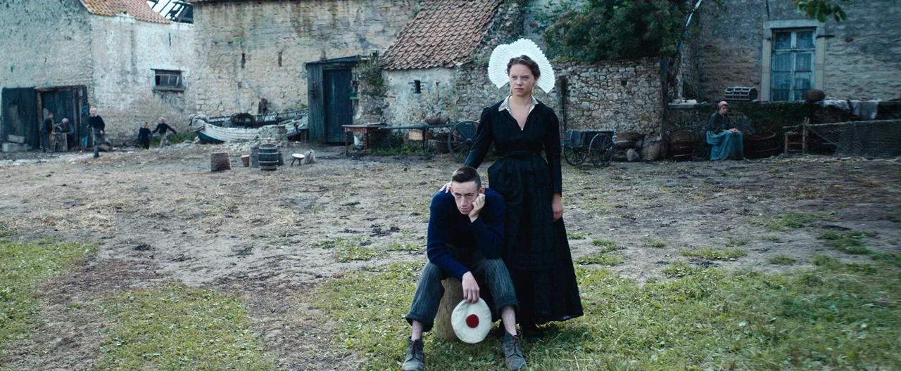 Slack Bay (2016) movie still: wide — A rural farmyard with stone outbuildings and muddy ground under an overcast sky. A young man sits on a wooden…