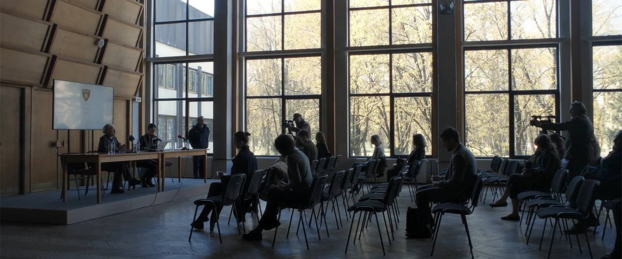 The Good Neighbor (2022) movie still: wide — A spacious conference hall features wood-paneled walls and floor-to-ceiling windows overlooking a park.…