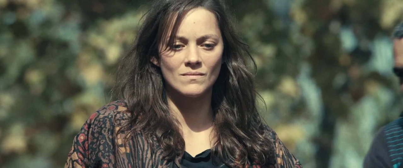 Rust And Bone (2012) movie still: medium — A woman stands in a park area with blurred trees in the background. She faces forward with her hair…