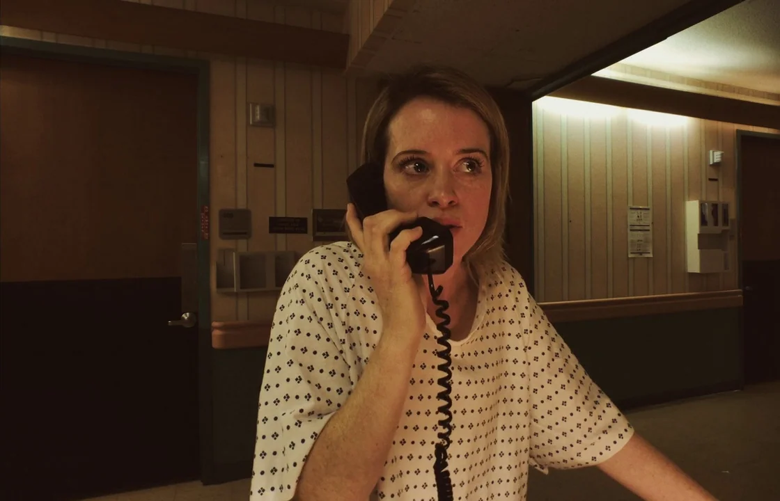 Unsane (2018) movie still: medium — A hospital hallway features institutional beige striped wallpaper and dark wooden doors. A woman wearing a…