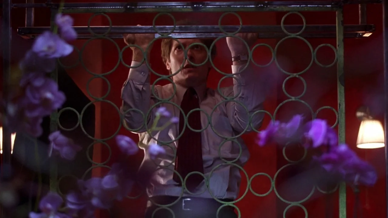 Secretary movie still: medium — A dimly lit room with walls painted a deep saturated red. A man grips the frame of an ornate green metal divider…