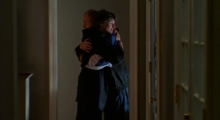 Schizopolis movie still: medium — A narrow home corridor features plain walls and a dark door frame. A man and a woman stand together in a tight…