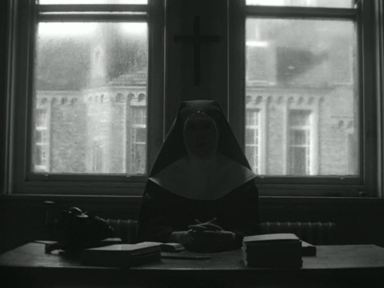 Death and Transfiguration (1983) movie still: medium — A dark office space with a window overlooking a brick building facade. A nun sits behind a…