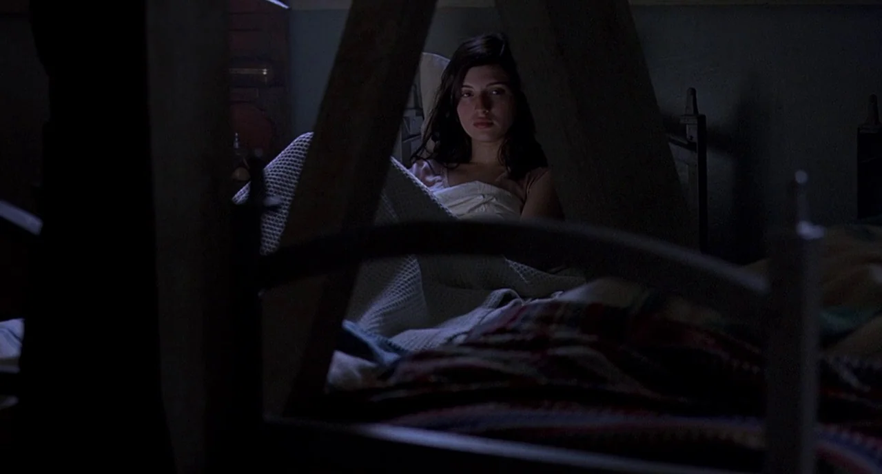 Cracks (2009) movie still: medium — A dimly lit room focuses on a person lying under a textured white blanket. Dark wooden slats of a bunk bed…