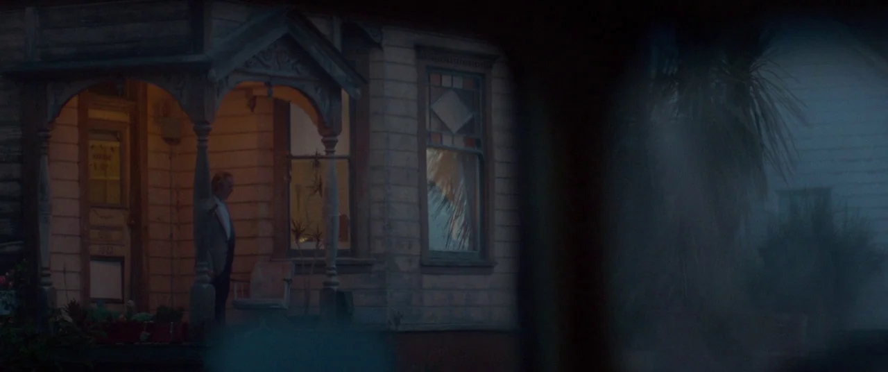 Woodshock (2017) movie still: wide — An aging wood-paneled house features a lit porch area with an ornate railing. An older man stands on the porch…