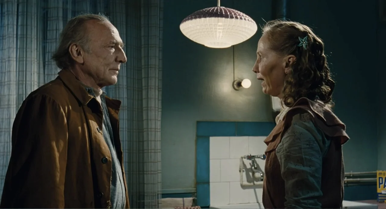Le Havre (2011) movie still: medium — A kitchen interior features tiled walls and a vintage pendant lamp hanging above. An elderly man and a woman…