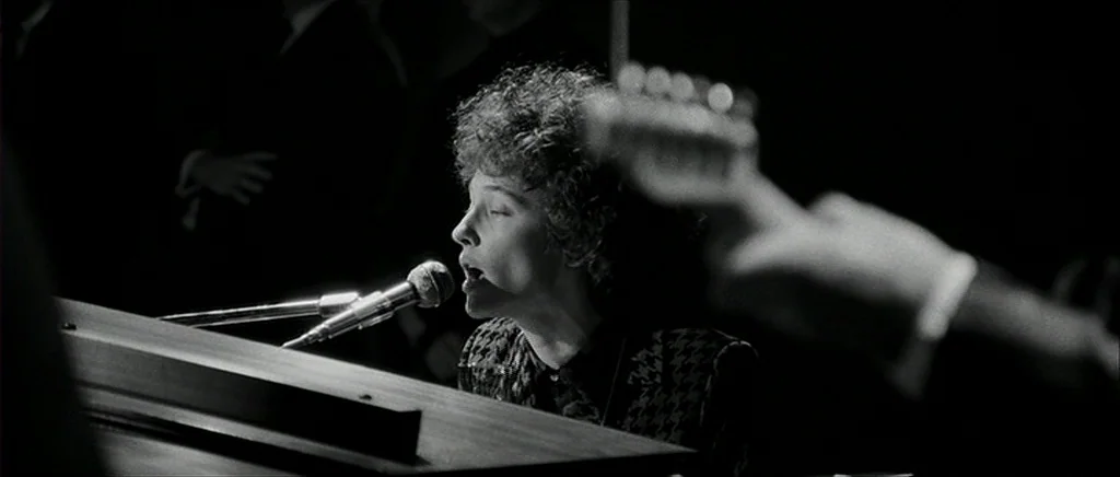 I’m Not There (2007) movie still: medium — A dark, dimly lit stage with a musician performing at a piano. The performer sings into a microphone…