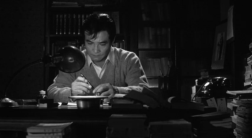 Mishima: A Life in Four Chapters (1985) movie still: medium — A cluttered dark study features shelves filled with books and stacks of paper. An…