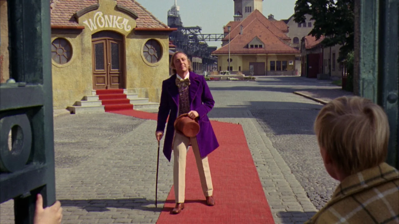 Willy Wonka & the Chocolate Factory (1971) movie still: fullbody — A man in a vibrant purple coat stands on a red carpet outdoors. He holds a top…