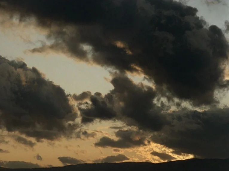 Hail Mary (1985) movie still: establishing — A dense layer of dark clouds fills the sky during a fading sunset. Faint warm light emanates from the…