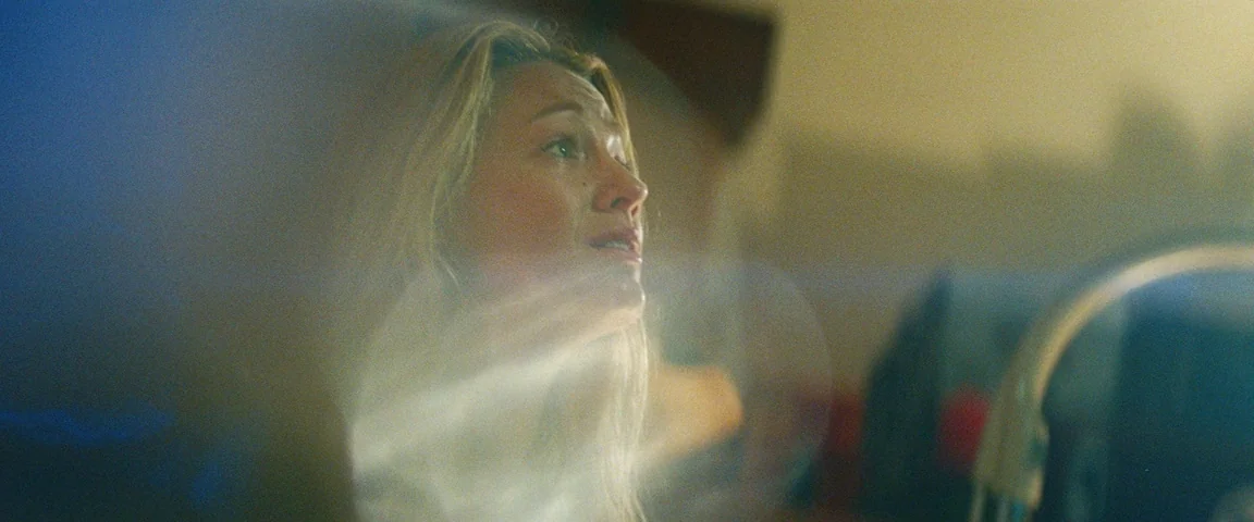 Savages (2012) movie still: closeup — A dimly lit interior space with soft, blurred light flares obscuring the foreground. A blonde woman looks…