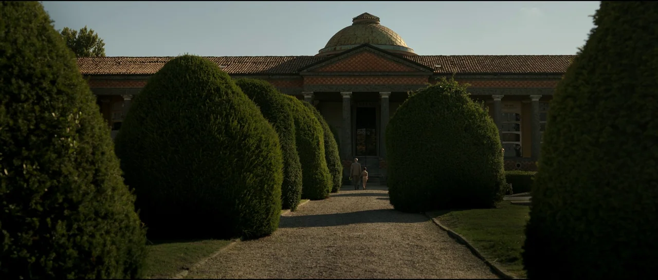 Ferrari (2023) movie still: wide — A stone building with a central dome sits at the end of a long path. An adult man and a small child walk together…