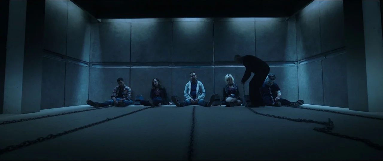Jigsaw (2017) movie still: wide — A windowless concrete bunker features modular panel walls and a cold blue aesthetic. Five people sit in a row on…