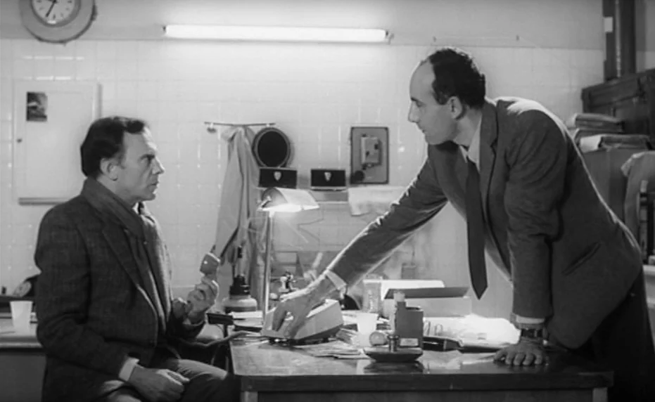 Vivement Dimanche (1983) movie still: medium — A cramped, tiled room contains a wooden desk covered with office supplies and paperwork. Two men…