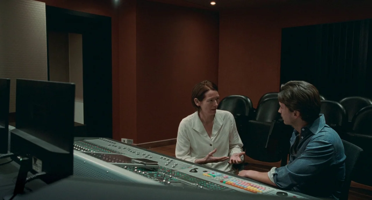 Memoria (2021) movie still: medium — A recording studio with red acoustic wall panels and rows of black chairs. A man and a woman sit at a large…