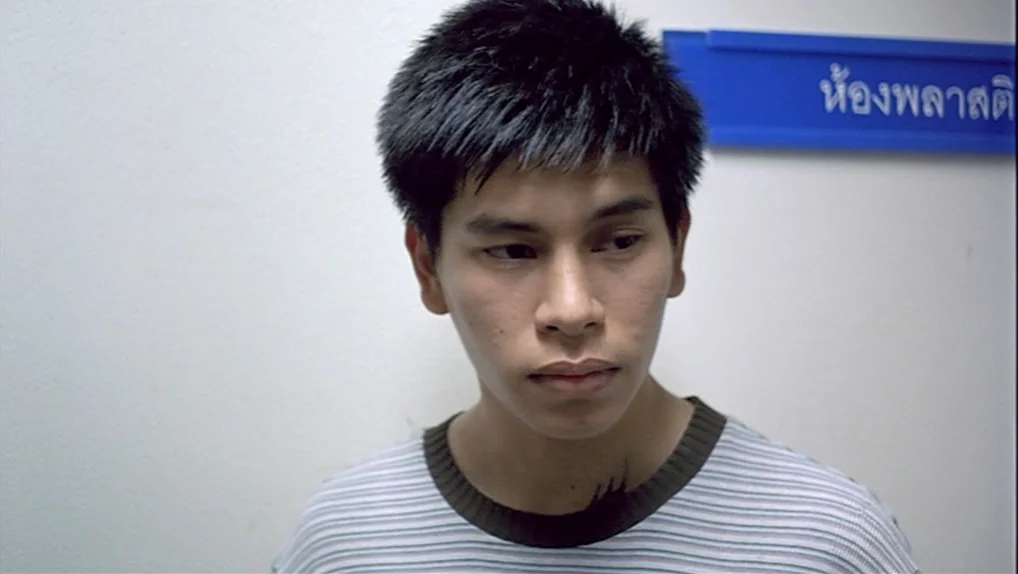 Syndromes and a Century (2006) movie still: closeup — A bright white interior wall displays a blue sign with Thai text. A young man stands in front…