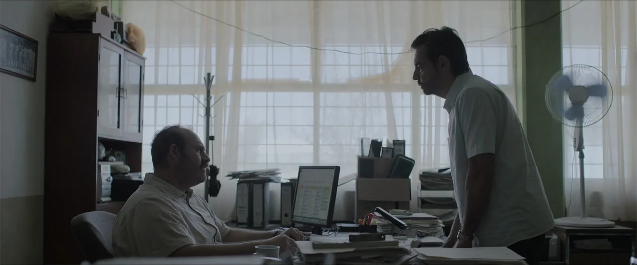 Radical (2023) movie still: medium — A cluttered office features a desk with a computer monitor, papers, and stacks of files. Two men engage in a…