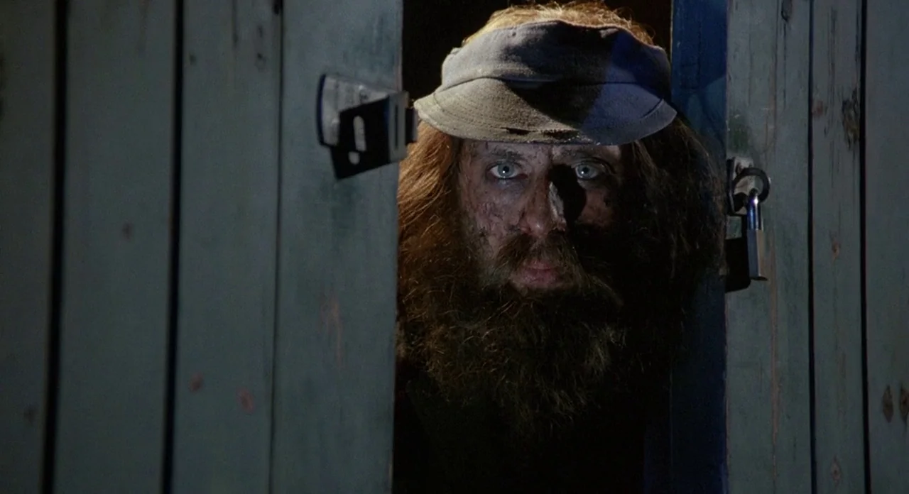 Hellraiser (1987) movie still: closeup — A man with a thick beard stands behind a dark, weathered wooden door gap. He looks forward with wide eyes…