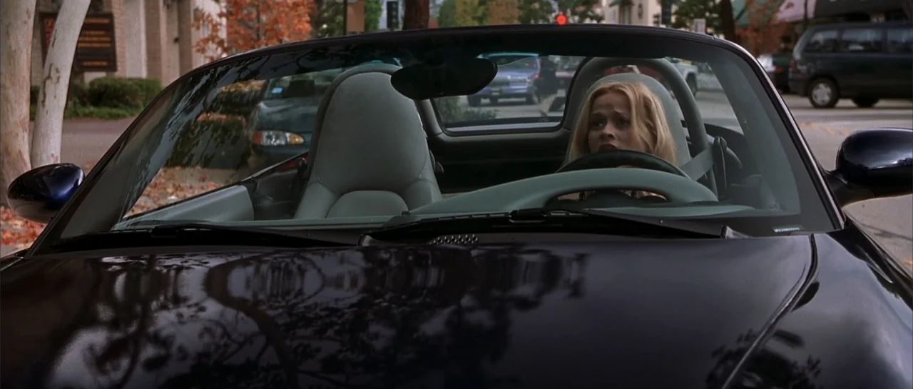 Legally Blonde (2001) movie still: medium — A dark convertible car is parked on a suburban street with mature trees visible. A blonde woman sits…