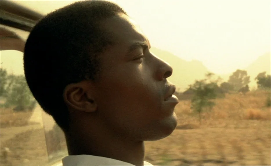 Chocolat (1988) movie still: closeup — A man rides in a vehicle past a dry, dusty field under golden light. He leans his head against the window…