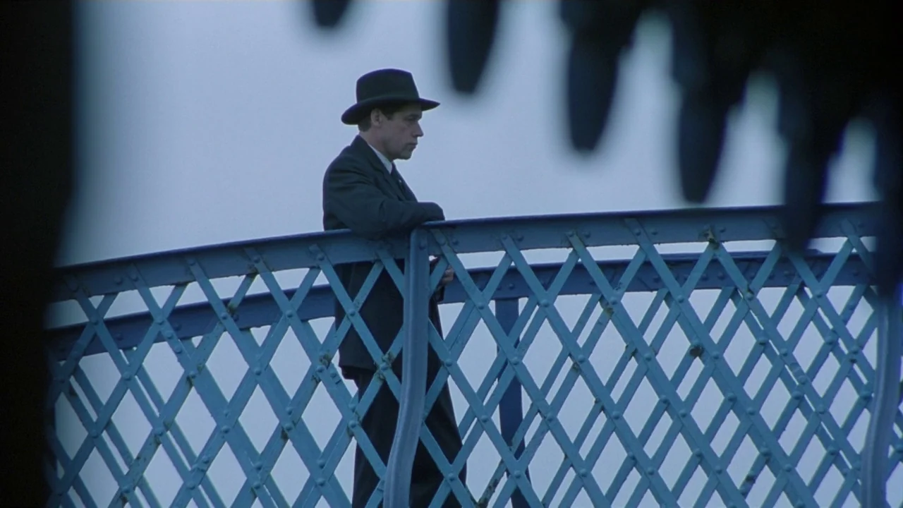 Michael Collins (1996) movie still: medium — A man in a dark suit stands on a bridge with blue latticework railing. The man looks forward while…