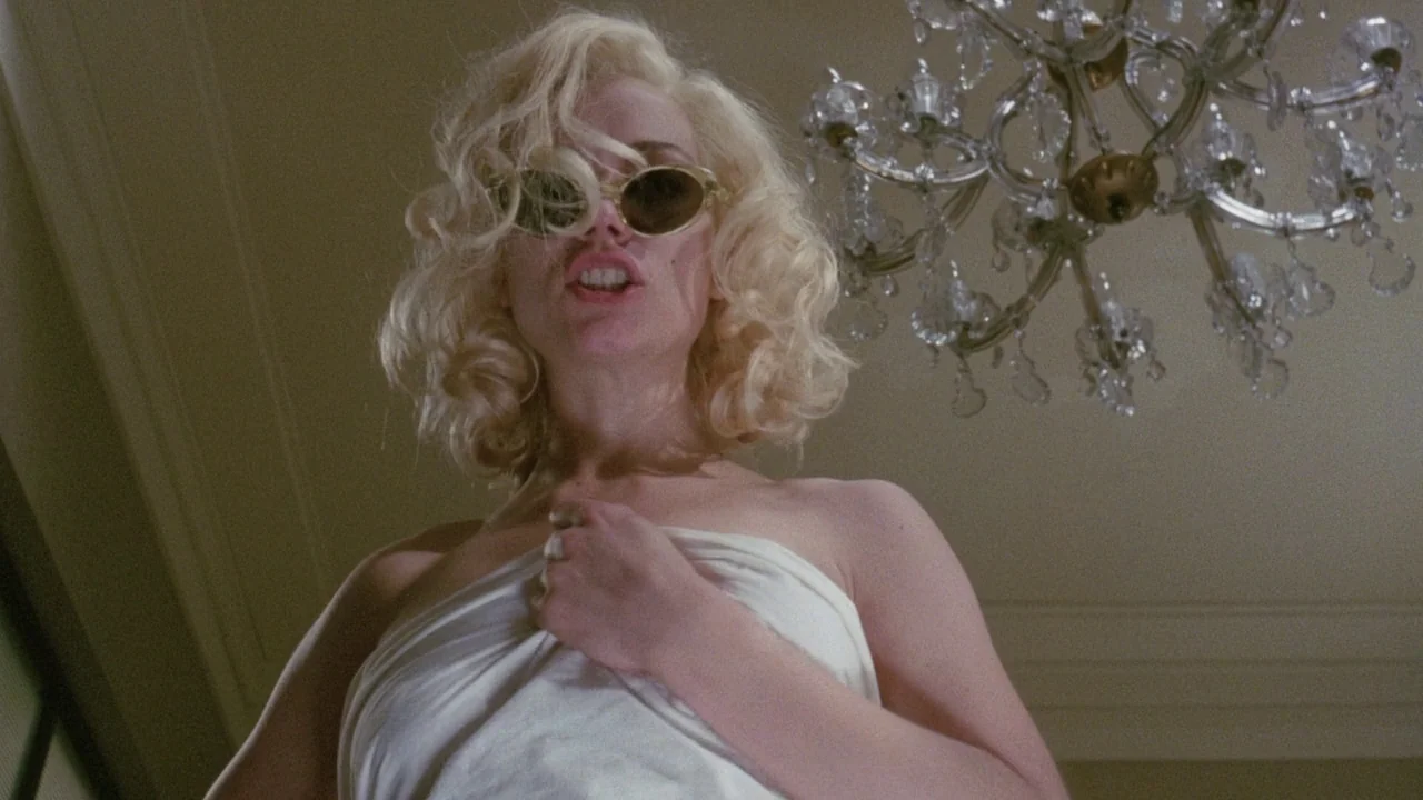 Insignificance (1985) movie still: medium — A room with a large overhead crystal chandelier and plain wall surfaces. A blonde woman wearing…