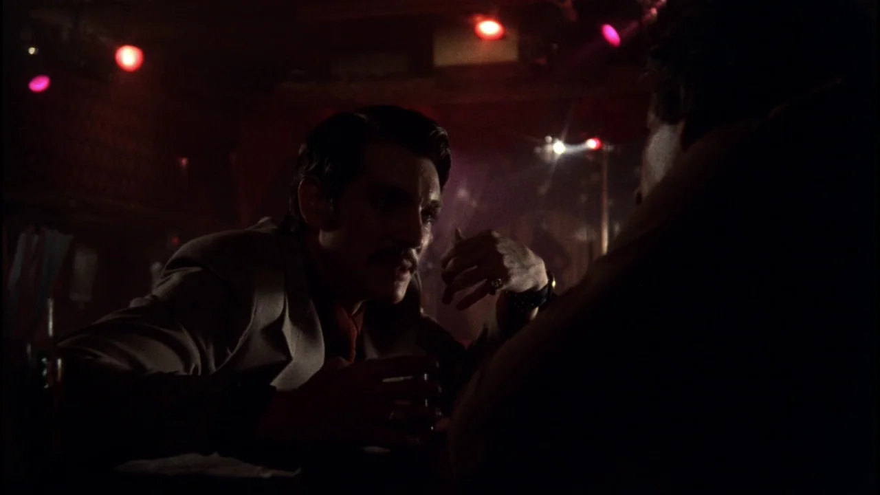 Star 80 (1983) movie still: medium — A dimly lit bar setting with red ambient lighting and visible glassware. A man in a suit engages in…
