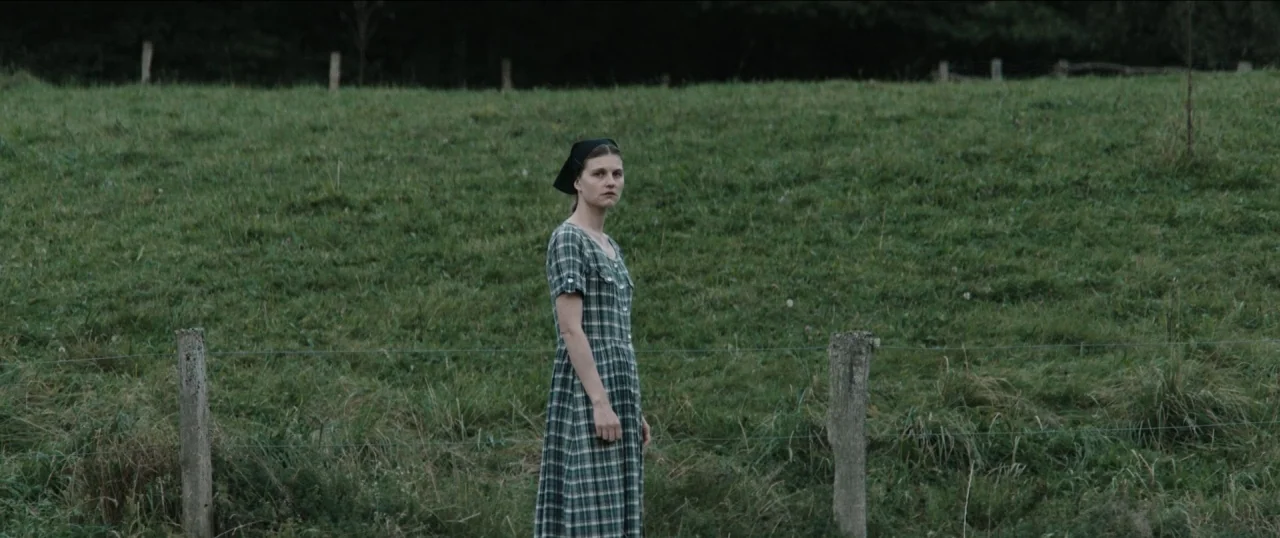Good Favour (2017) movie still: fullbody — A rural pasture with lush green grass and a wooden fence running horizontally. A woman in a plaid dress…