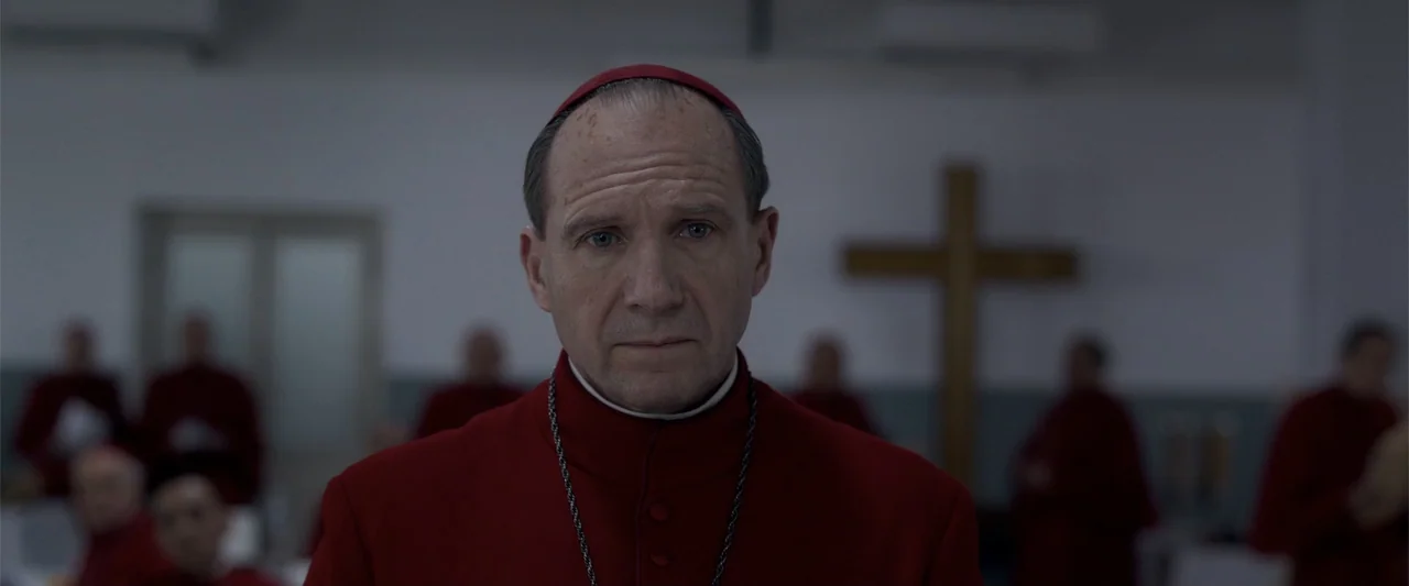 Conclave (2024) movie still: closeup — A man wearing a cardinal's red cassock stands in a plain, sterile white-walled interior. Other individuals in…