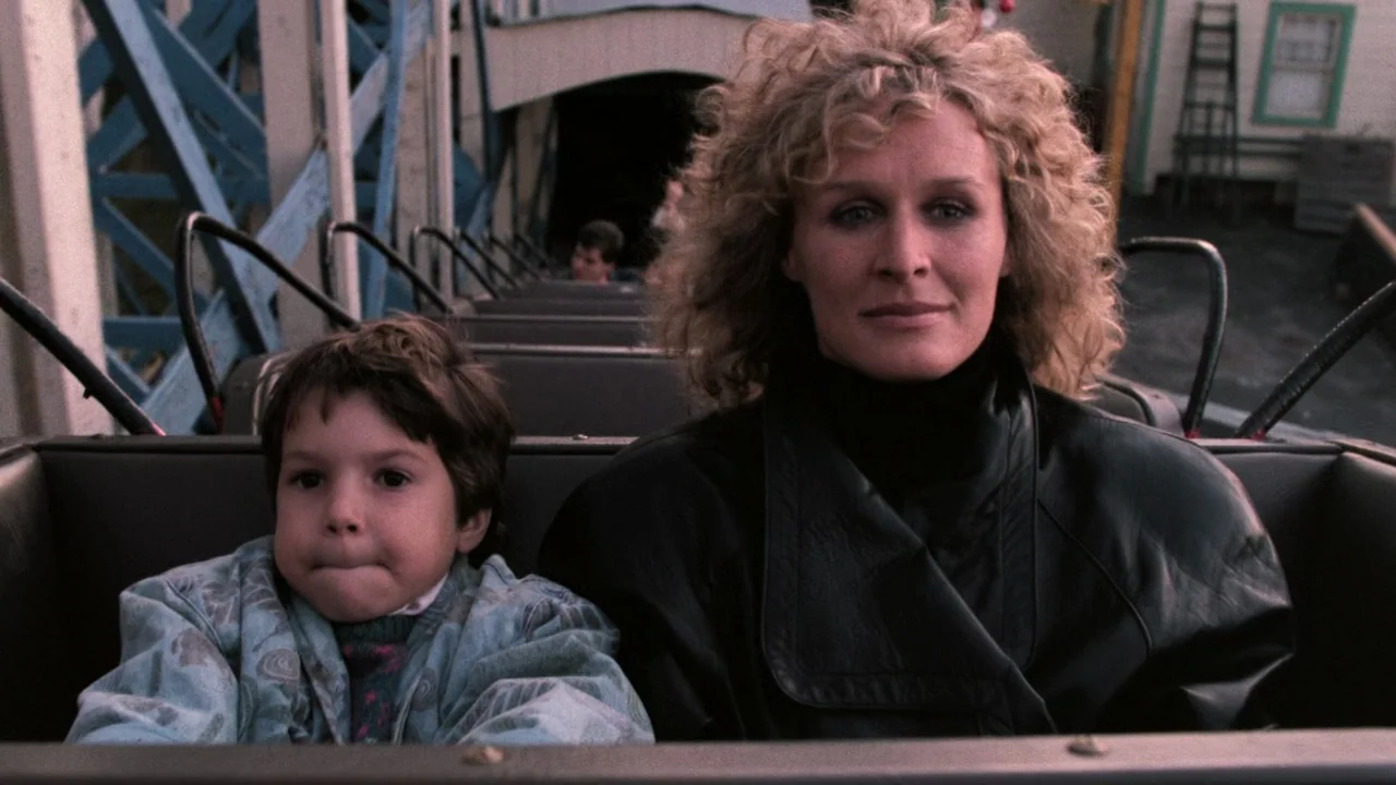 Fatal Attraction (1987) movie still: medium — A woman and a small boy sit inside a roller coaster car. The amusement park structure is visible in…