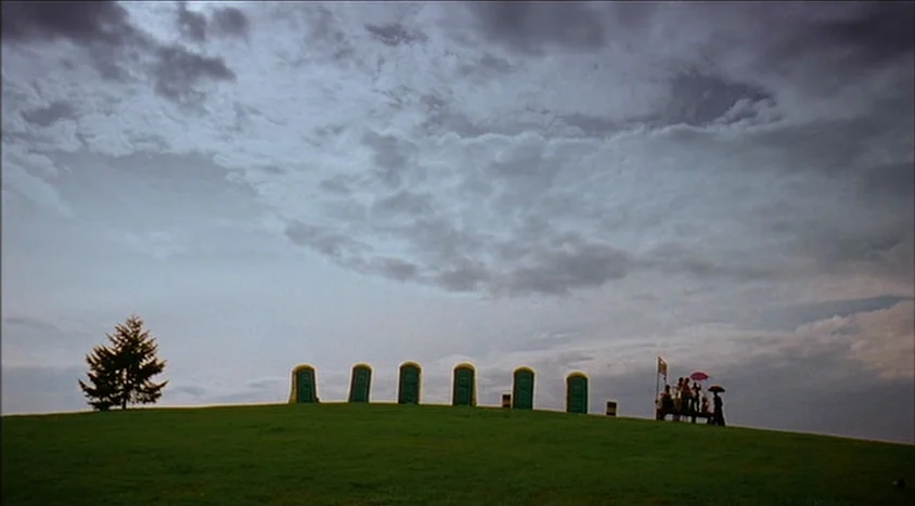 Hedwig and the Angry Inch (2001) movie still: wide — A green grassy hill stretches toward a horizon under a dense, cloudy sky. A row of six portable…