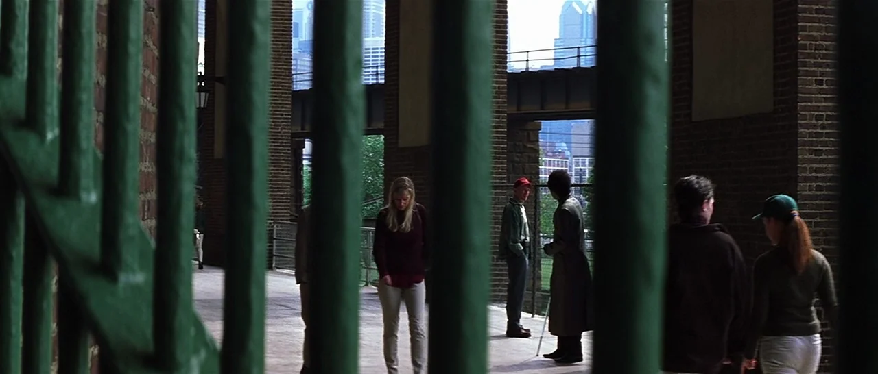 Unbreakable (2000) movie still: wide — A stone and brick pedestrian area beneath an elevated rail structure is visible. Several individuals stand…