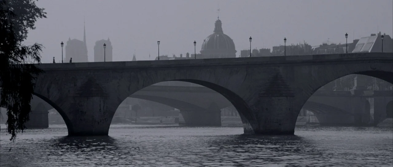 Angel-A (2005) movie still: establishing — A stone arched bridge spans across a wide, flowing river in the distance. The cityscape includes a…